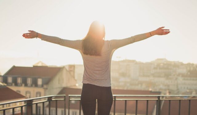 Güneşli balkonda kollarını açarak sabah ışığını karşılayan kadın, sağlıklı hayatın keyfini yaşıyor.