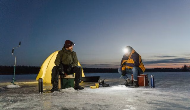 Karla kaplı gölde çadırın önünde balık tutmak için bekleyen iki adam, kış akşamı macerası yaşıyor.