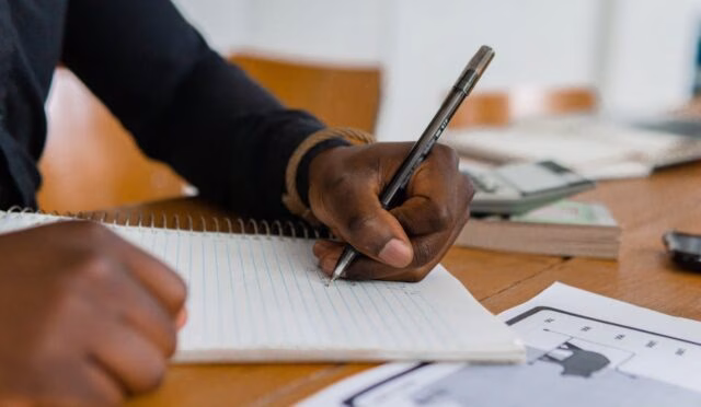 Eğitimin önemi üzerine notlar alan bir el, ahşap masa ve kalem ile yazma anını yakalıyor.